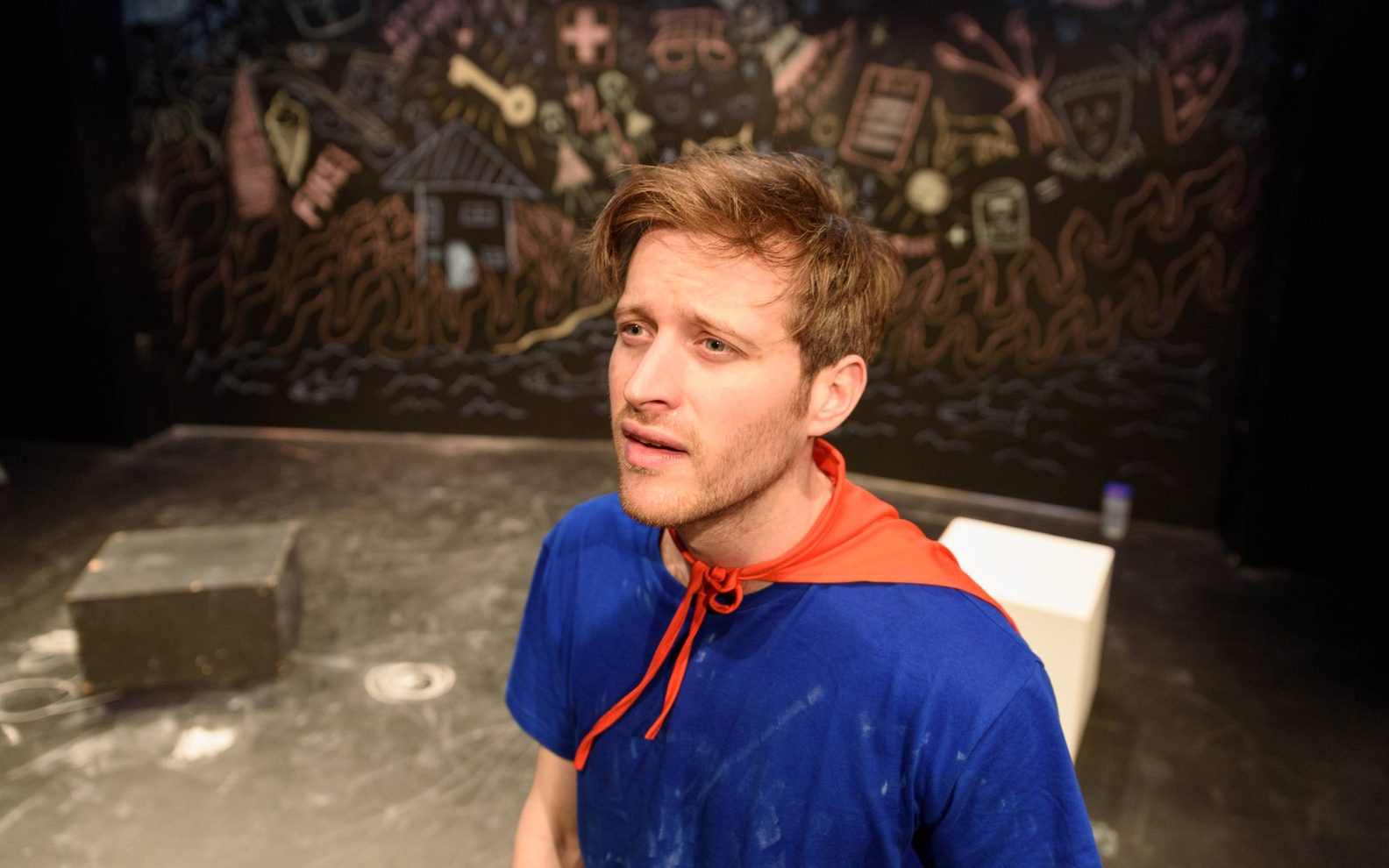 A man wearing a blue t-shirt and red cape. The wall behind him is covered in chalk drawings.