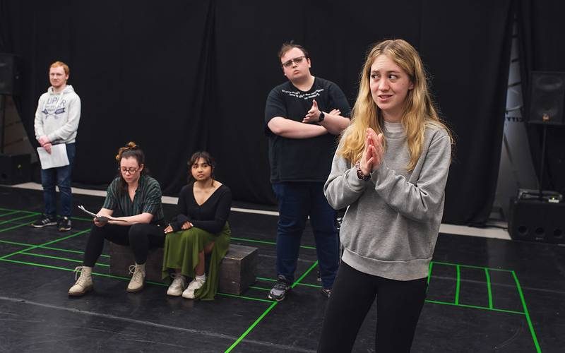 52827669280_6c6f4f24fe_c-aspect-ratio-1600-1000 Several young people rehearse a performance in an empty, all-black space. There are lines of bright green tape on the floor to mark positions.