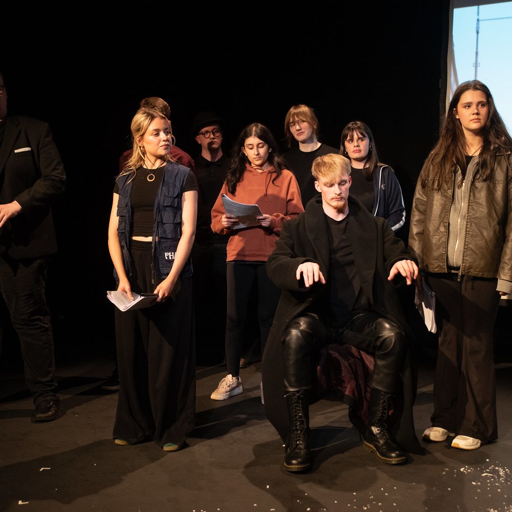 A group of young people stand on a small stage looking in the same direction. One of them is miming playing the piano.