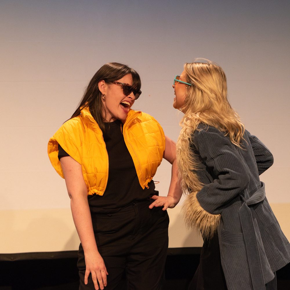 Two young women smiling broadly at each other while performing on stage. One is wearing sunglasses and a bright yellow puffer gilet.