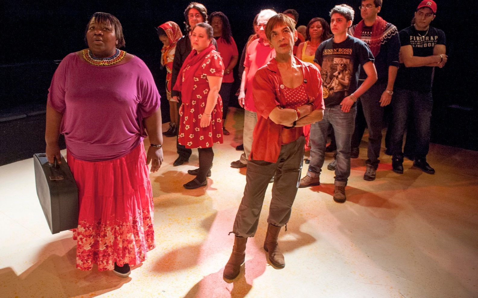 An ensemble cast. A group of people wearing bright clothing standing together. One person is holding a suitcase.