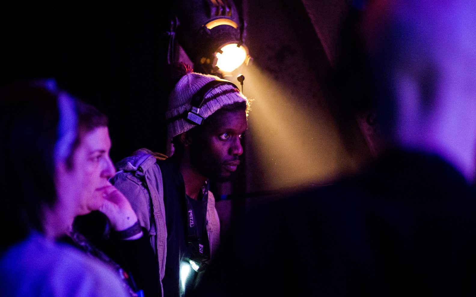 Barry Forde standing in the wings. He is wearing a headset and a torch around his neck. There is a spotlight above him.