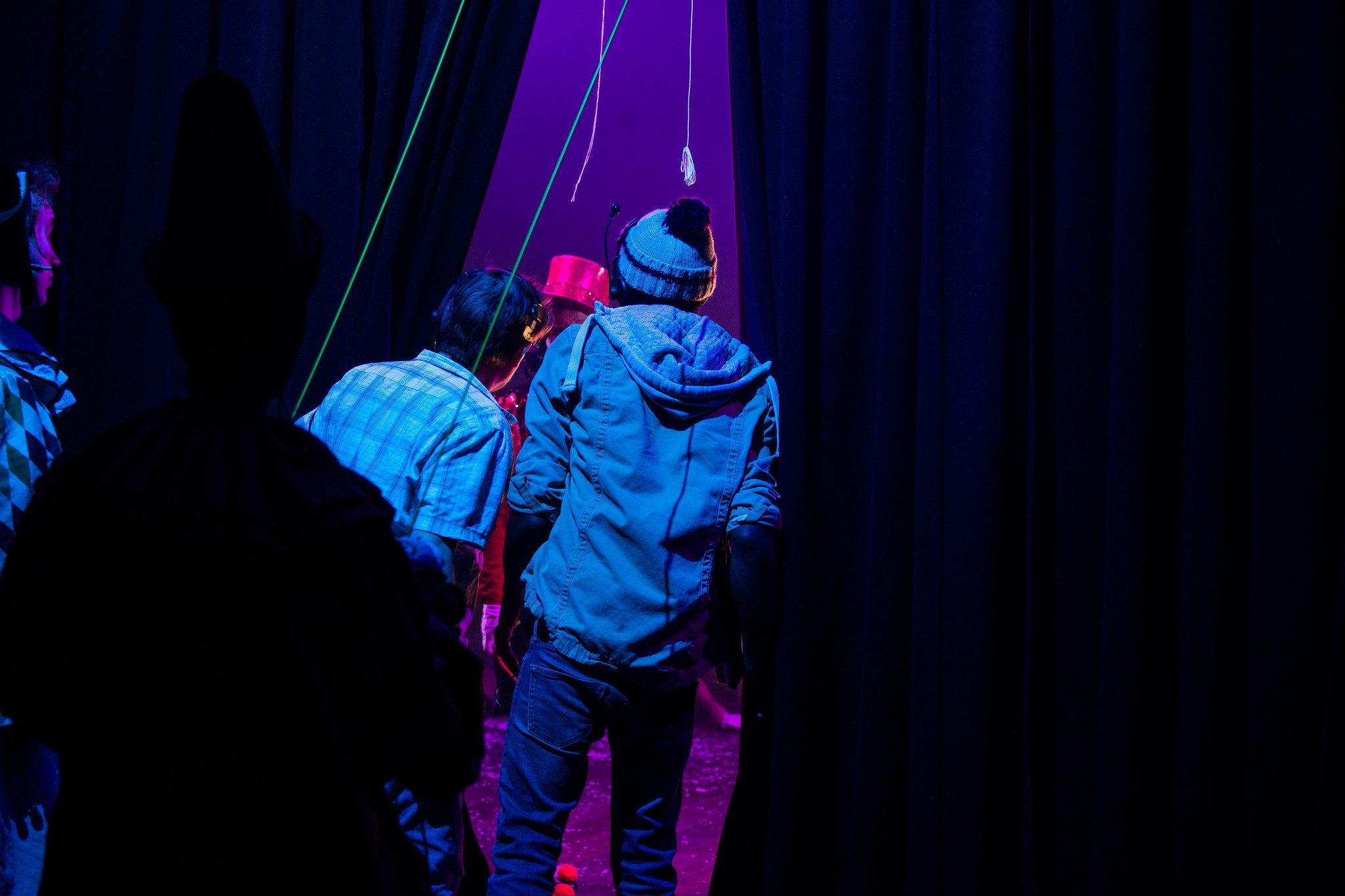 Stage managers and actors standing backstage. They have their backs to the camera and are looking through some theatre curtains.