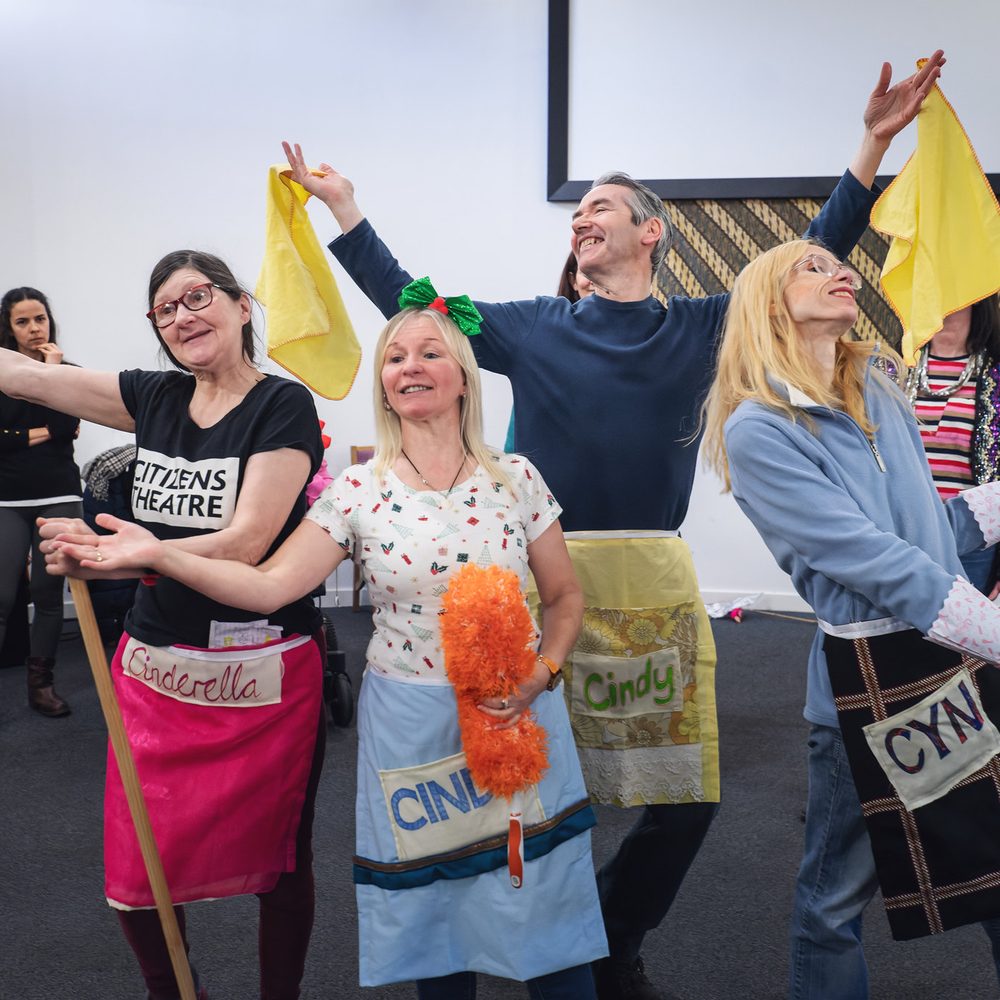 Four people rehearsing a production. They are wearing aprons and carrying cleaning implements.