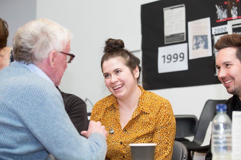 Carly McCaig speaking to a participant and smiling broadly. There is a cup of tea on the table in front of her.