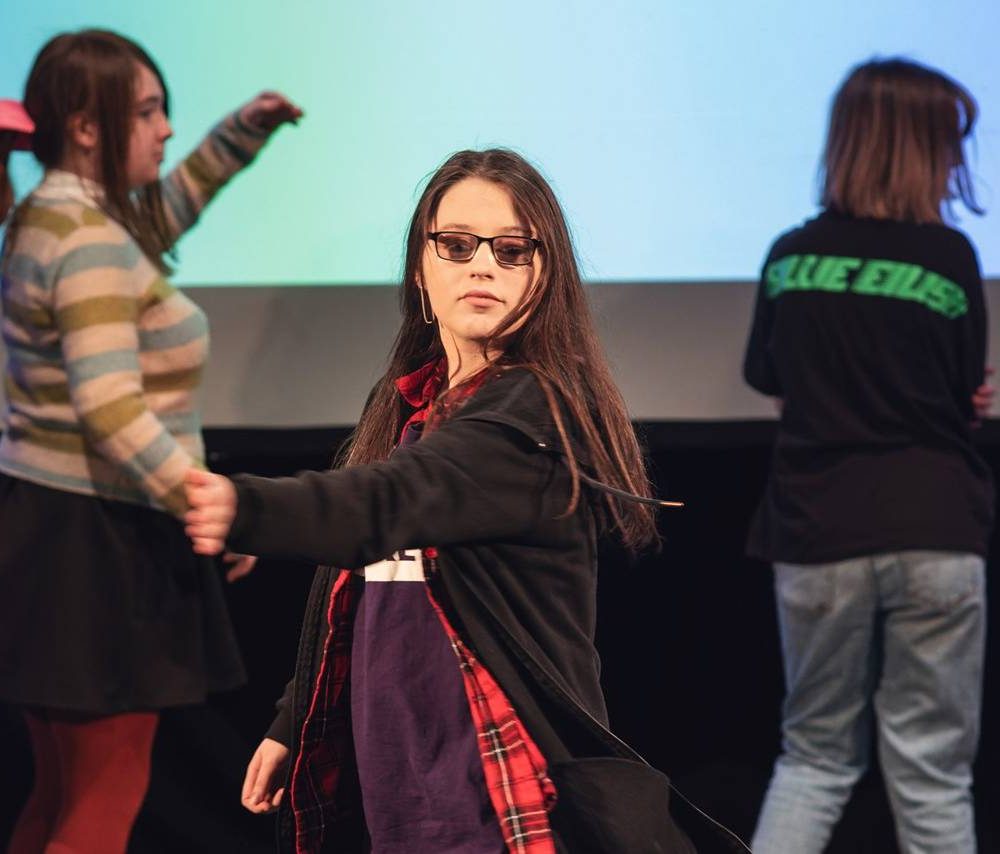 A young woman is dancing in the middle of a stage. There are two other people dancing behind her. A screen behind them says 'This song makes me feel seen.'