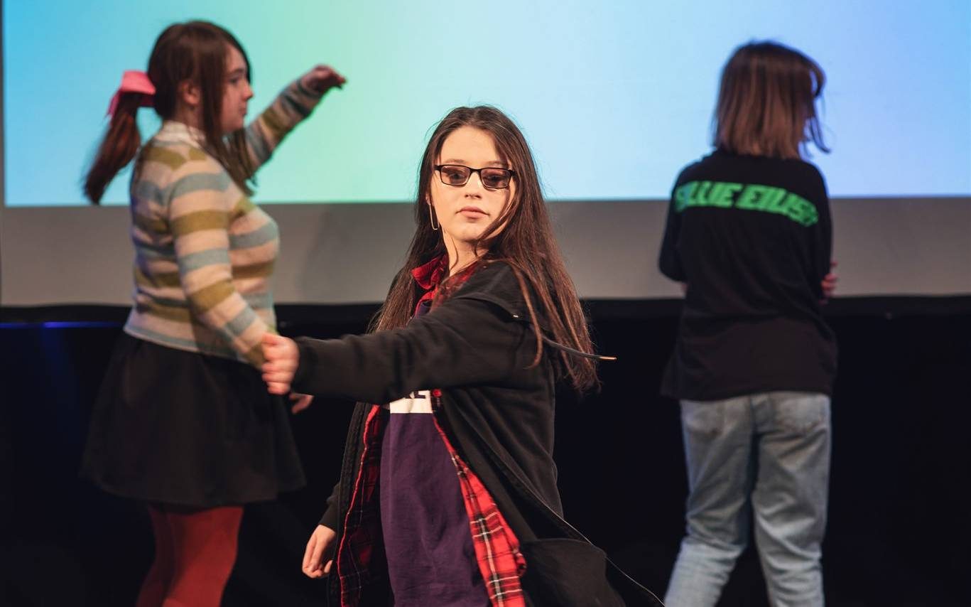 A young woman is dancing in the middle of a stage. There are two other people dancing behind her. A screen behind them says 'This song makes me feel seen.'
