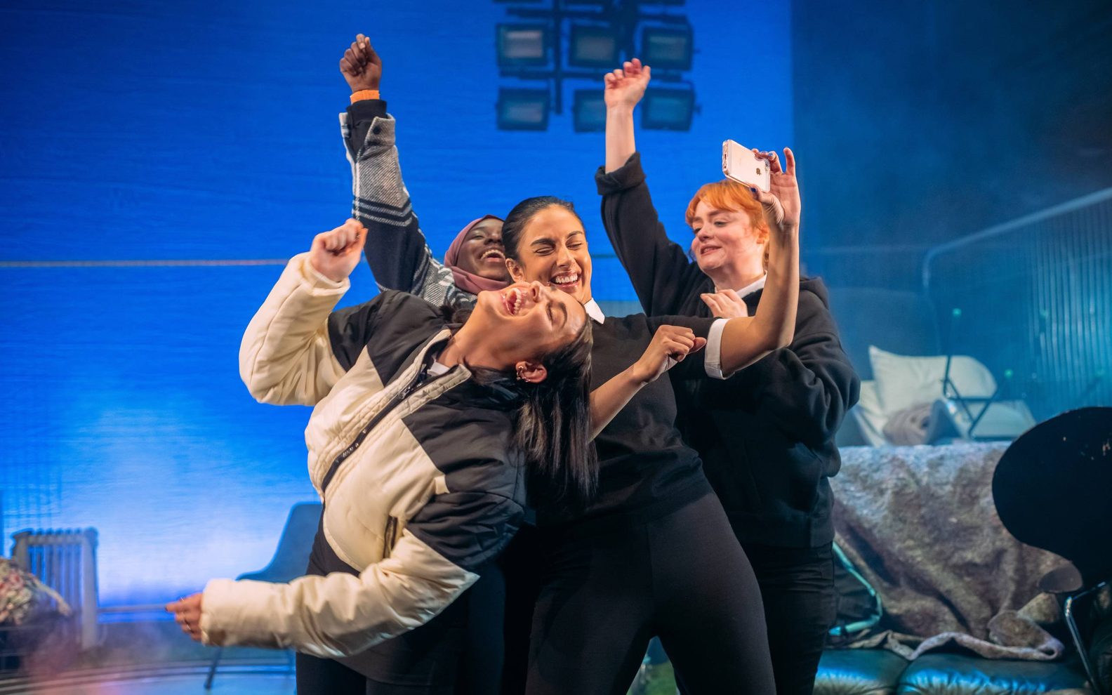 A stage show. Four women dressed in school uniform dancing and laughing together. One of them is taking a selfie of the group with her mobile phone. 