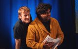 Two young men standing close together in front of a blue curtain. They both have expressive faces, and one is holding a script in his hand.