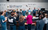 A group of actors in a rehearsal room. They are leaning over a long table.