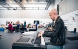 A man playing an electric piano. Behind him you can see a rehearsal room full of people.
