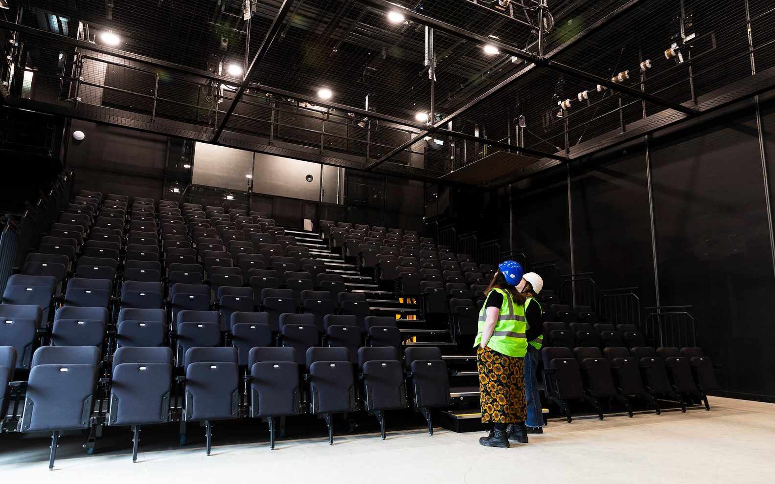 54380085743_de5803d9b8_k Two people standing in a a studio theatre space looking up towards the top of the seating bank. They are wearing high vis vests and hard hats