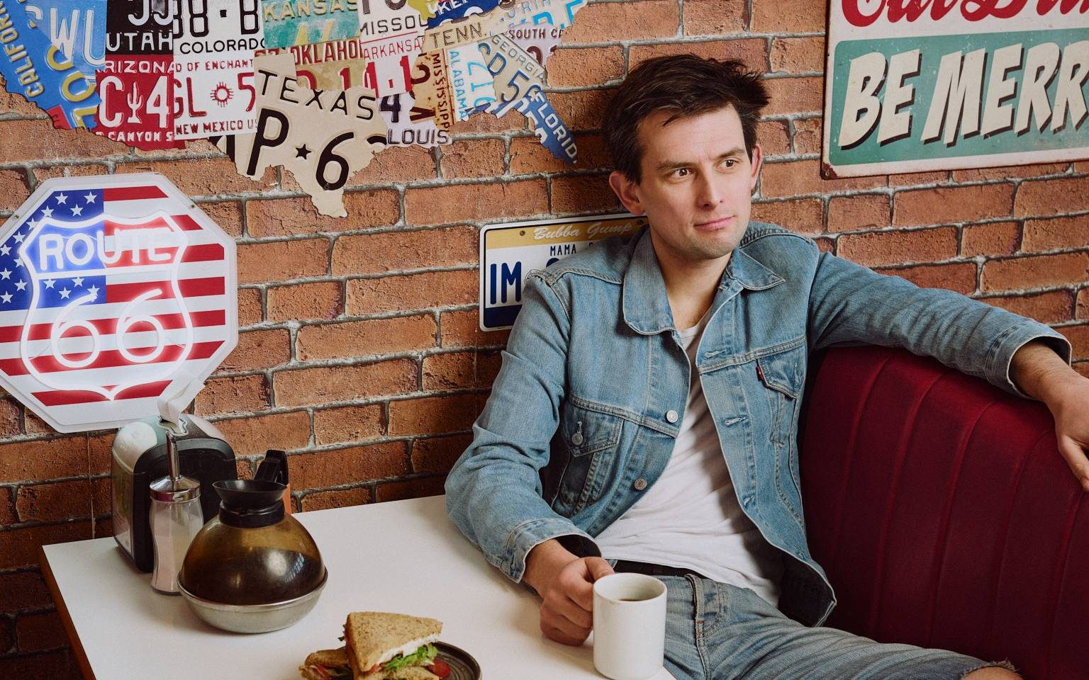 KIERAN-HODGSON-KH_0975-WEB-RES-COPYRIGHT-MATT-STRONGE-1-aspect-ratio-1600-1000 A man sitting at a dinner table, he's wearing all denim. It has an American feel.