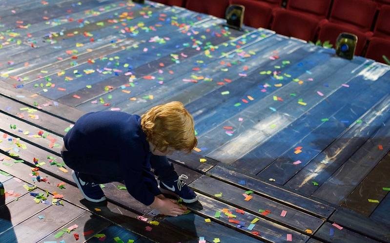 A young boy is crouched down on a stage. He is picking up small bits of brightly coloured pieces of confetti.