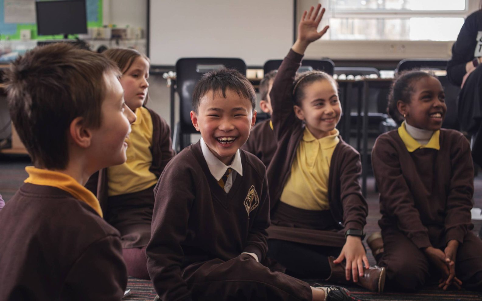 A group of primary pupils sat cross legged on the classroom floor. They are wearing brown and yellow school uniforms and are smiling broadly. One pupil has their arm raised in the air to answer a question.