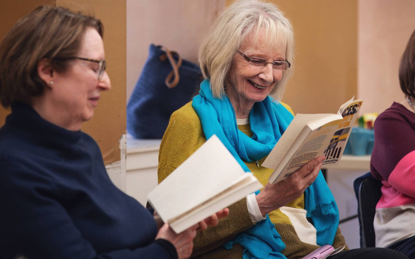 A play reading session. A woman with glasses and a bright blue scarf smiles while reading from a book.
