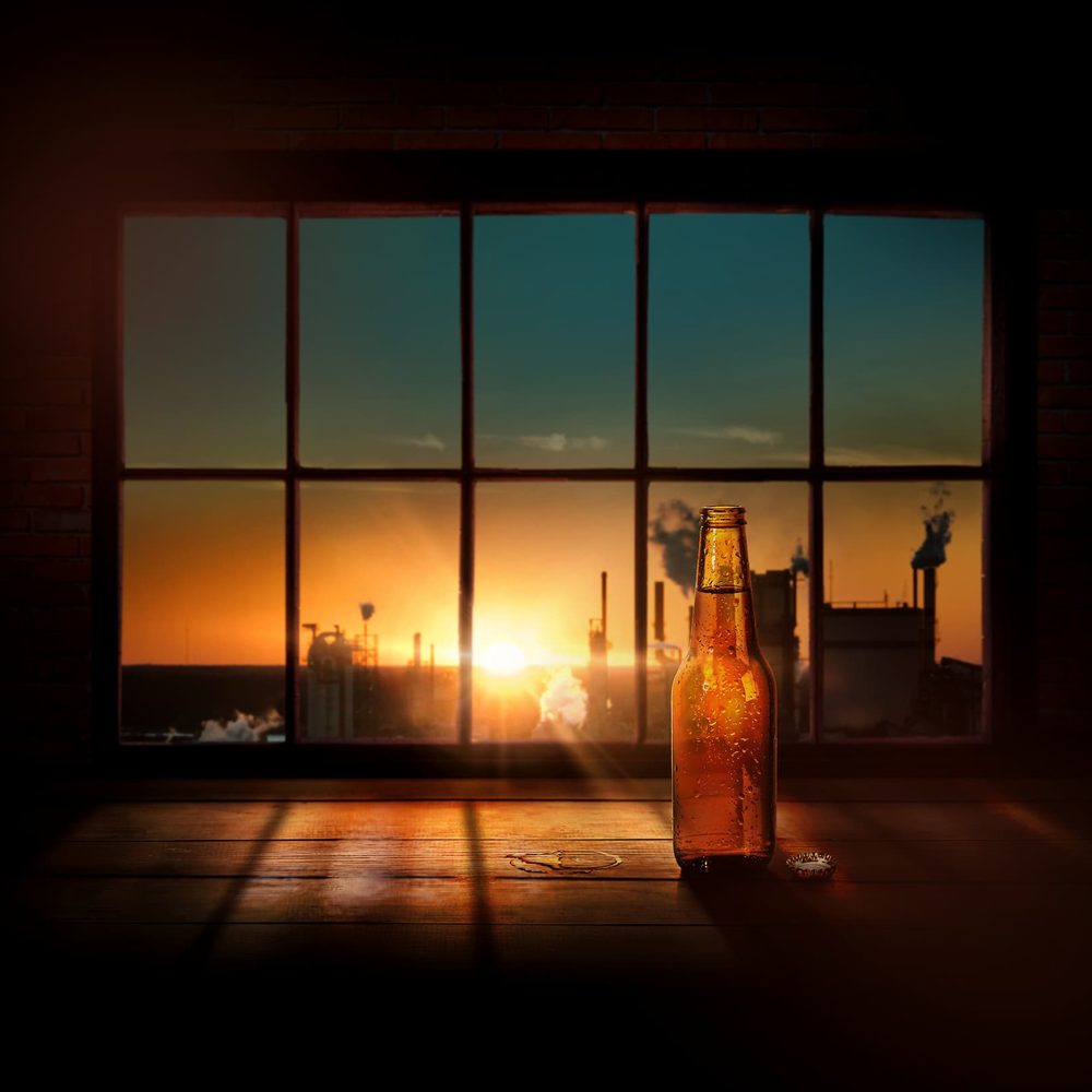 A brown beer bottle with condensation on a wooden bar, with a large window behind showing a sunset over a factory.