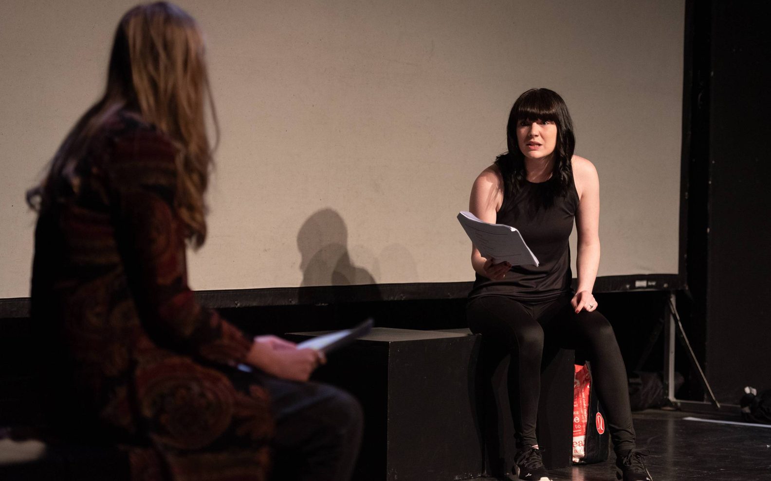A performance image. Two actors sit on stage talking to each other.