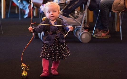 A young toddler is walking towards the camera. She is holding a bunch of red and yellow balloons. Three adults are in the background smiling at her.