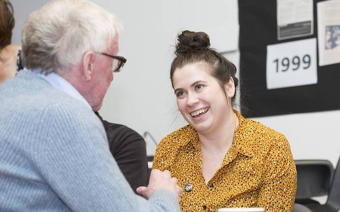 Carly McCaig speaking to a participant and smiling broadly. There is a cup of tea on the table in front of her.