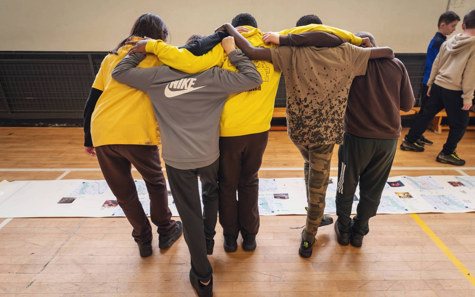 A group of primary pupils standing arm in arm with their backs to the camera. They are looking at a large collage on the ground.