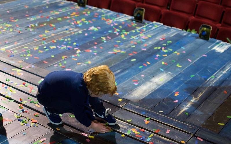 A young boy is crouched down on a stage. He is picking up small bits of brightly coloured pieces of confetti.
