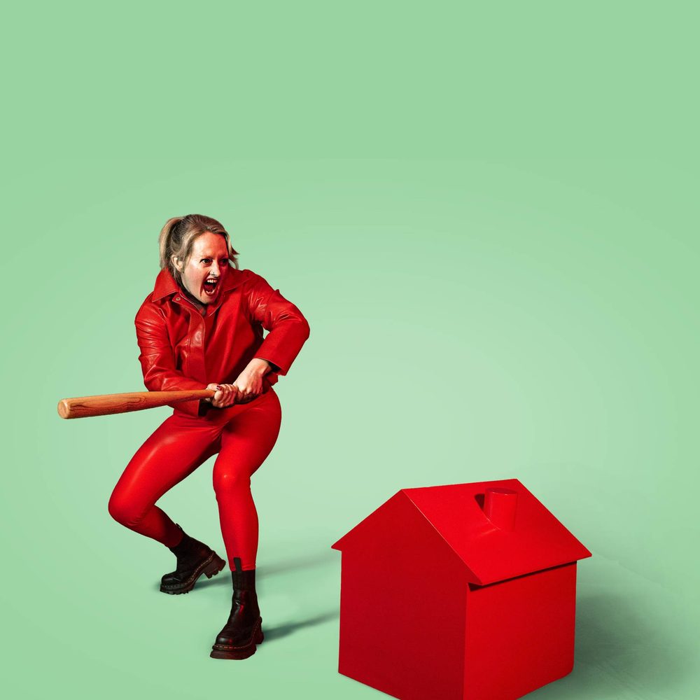 A woman dressed all in red with black boots holds a baseball bat. She is threatening to hit a small red house shaped like the Monopoly house piece.