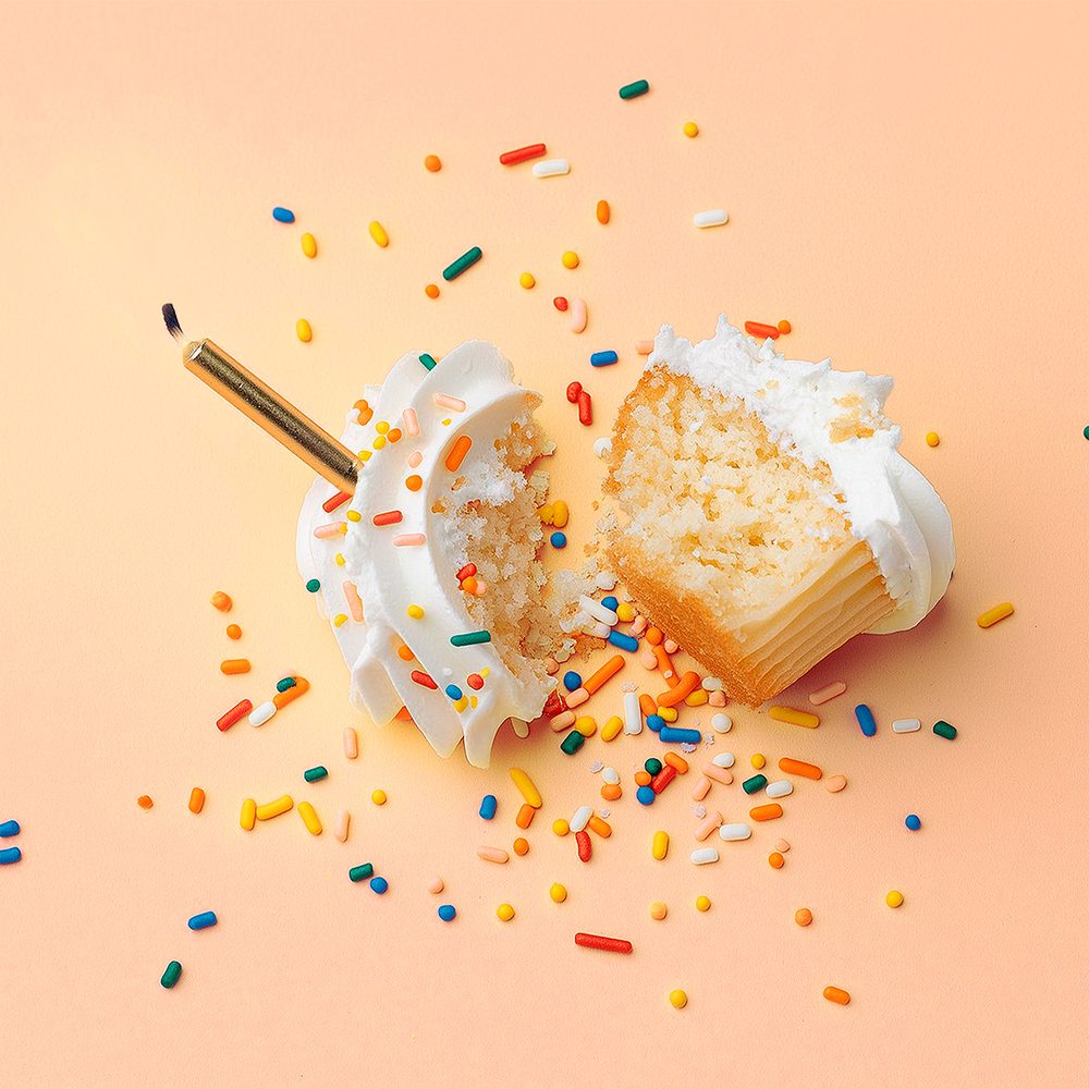 A cupcake with thick white icing and colourful sprinkles dropped onto the floor. The cake is broken in half and the sprinkles are scattered. There is a burnt out gold candle on the top of the cake.