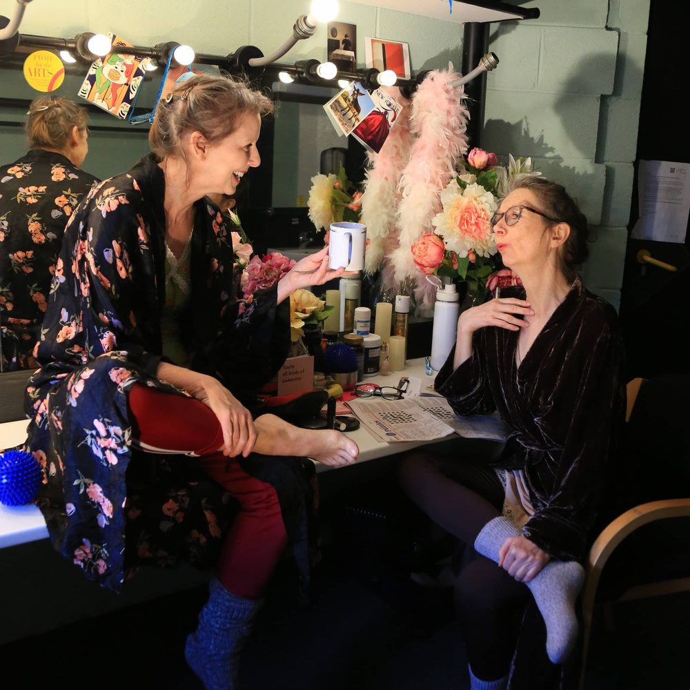 Two women sit in a busy dressing room, smiling.