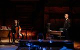 Two performers on stage. One is standing by a desk, hand in pockets. The other is sitting on a chair playing a cello.