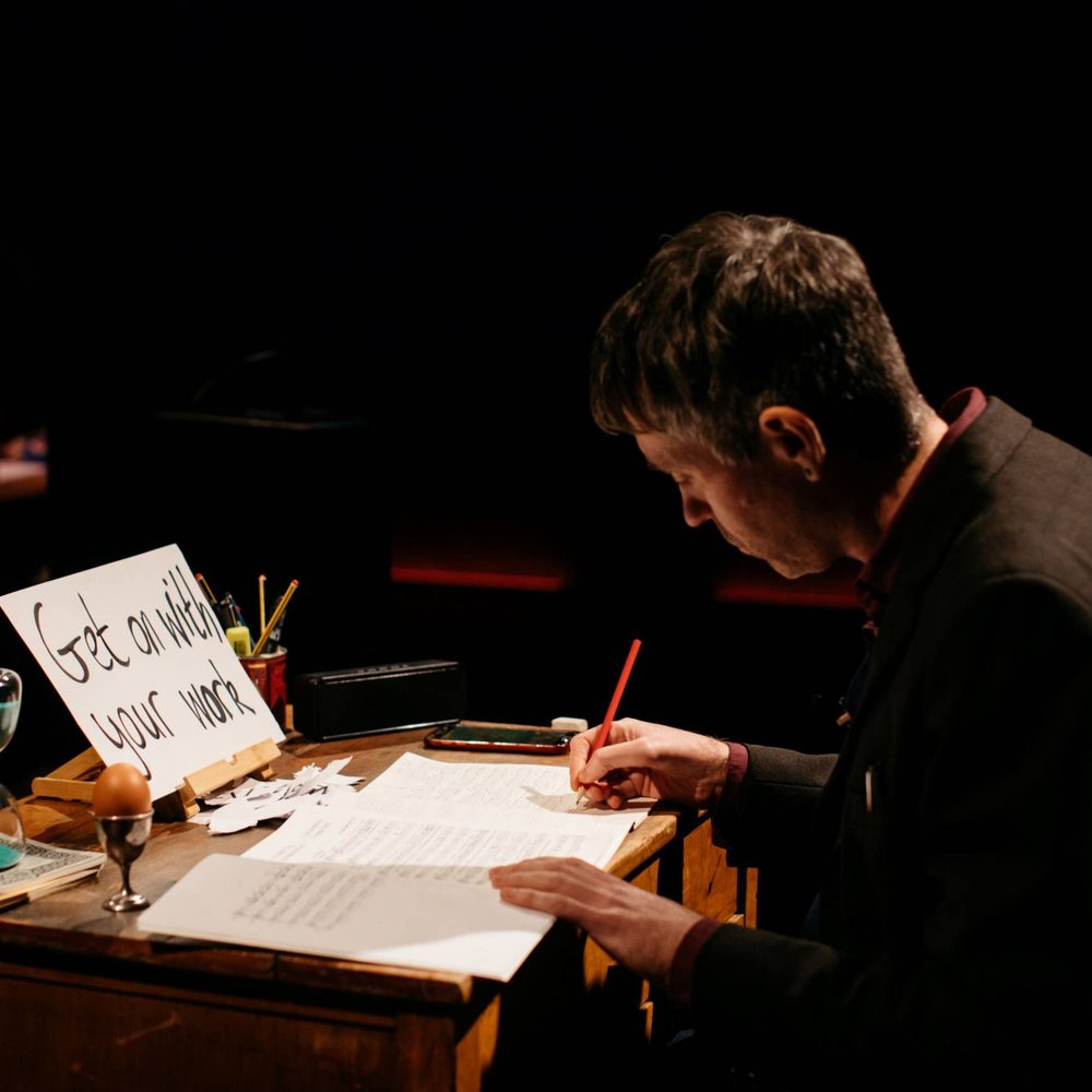A performer on stage. The man is sitting at a desk writing. On the desk is an egg in a egg cup, an hourglass timer and a mobile phone.
