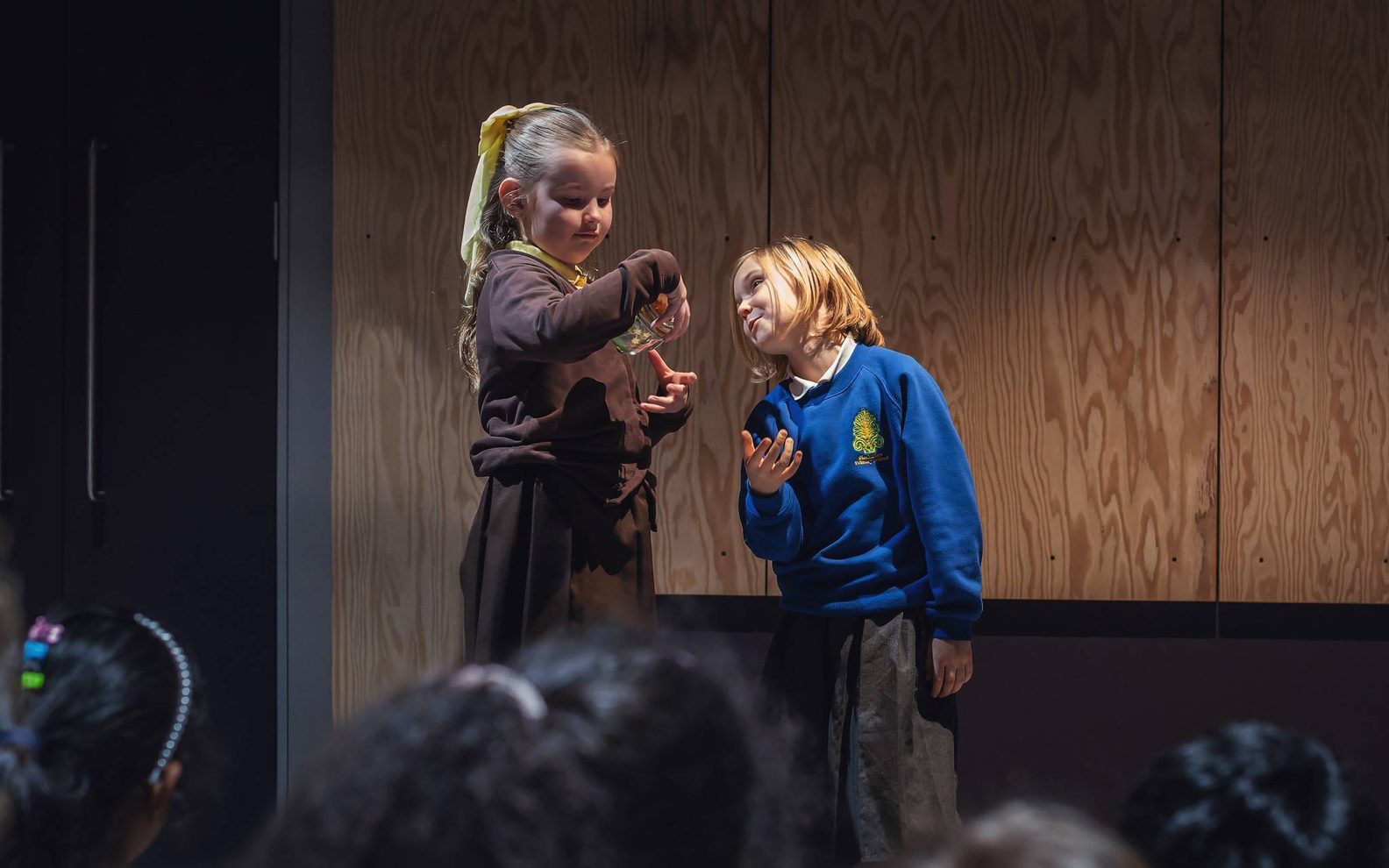 Two children, one in a brown uniform, and one in a blue performing in front of a group. One is holding a jar.