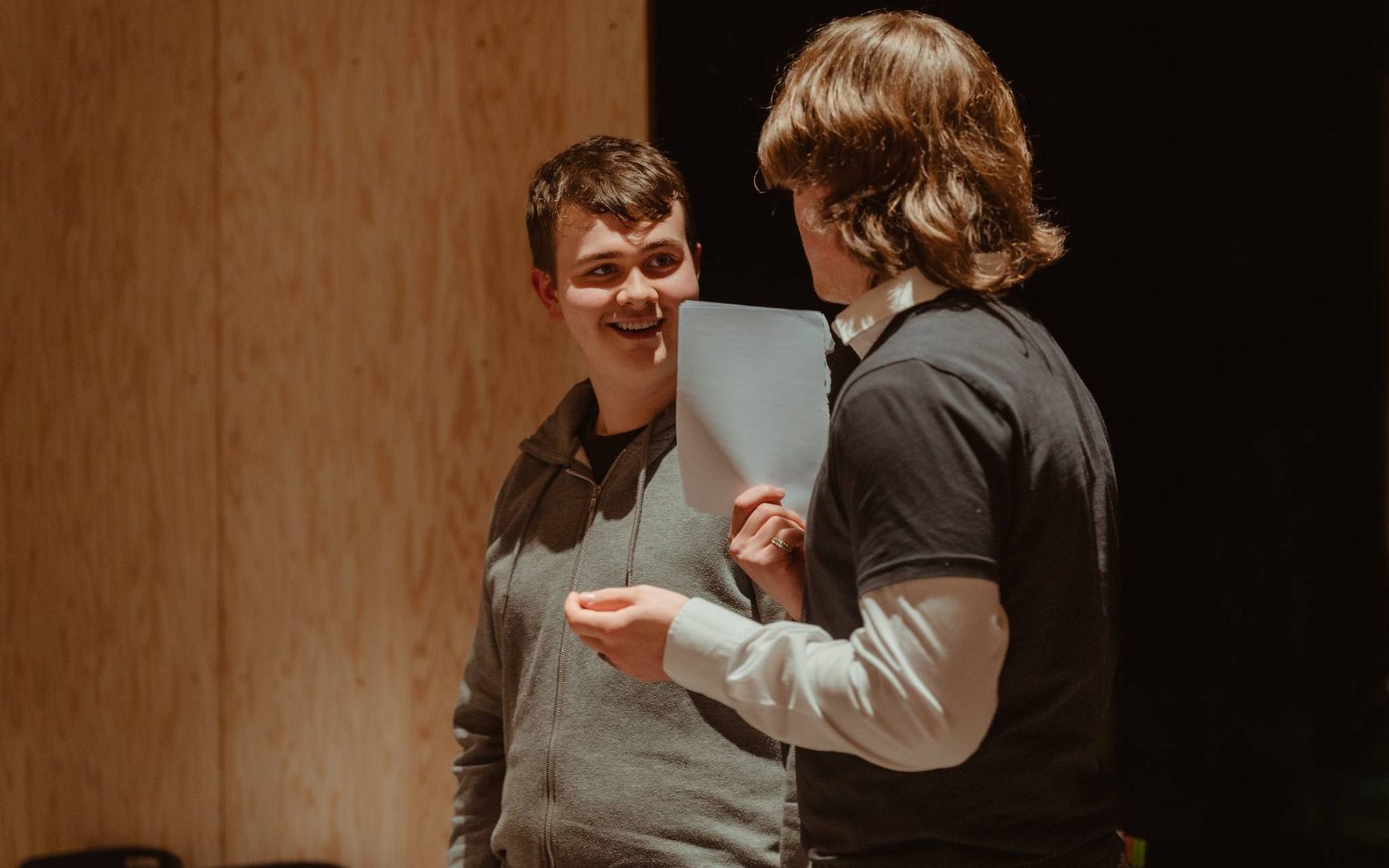 Two teenage boys chat to each other. One is holding a piece of paper.