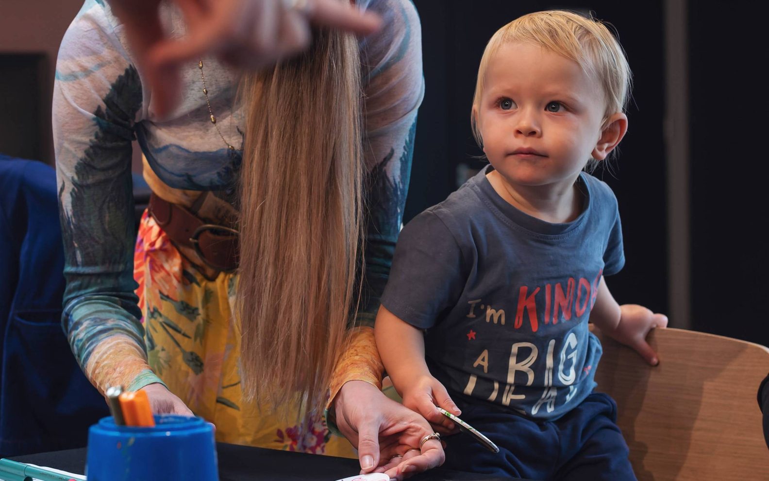 A table with craft supplies, with a woman and a toddler standing alongside it. The toddler is standing on a chair.