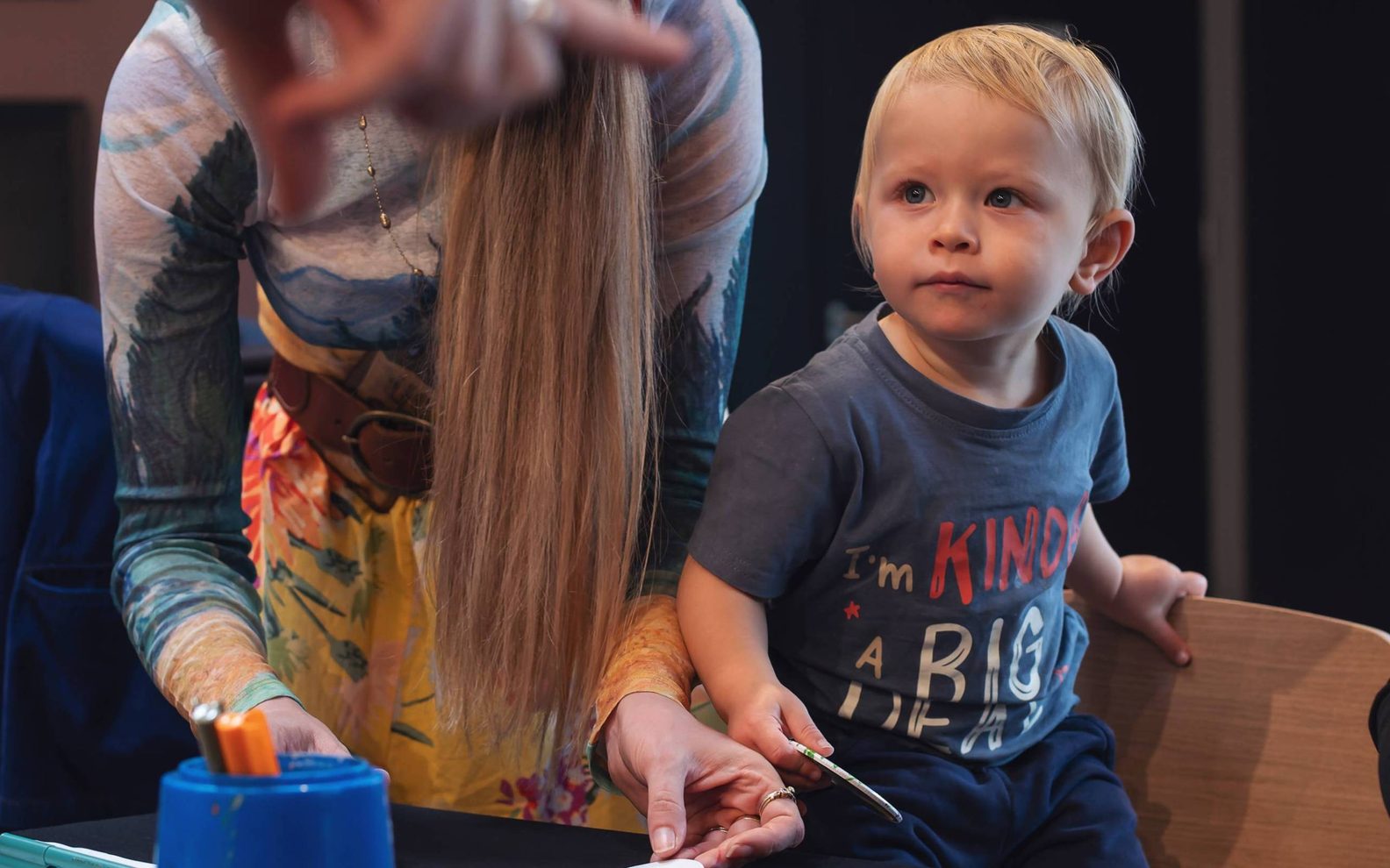 A table with craft supplies, with a woman and a toddler standing alongside it. The toddler is standing on a chair.